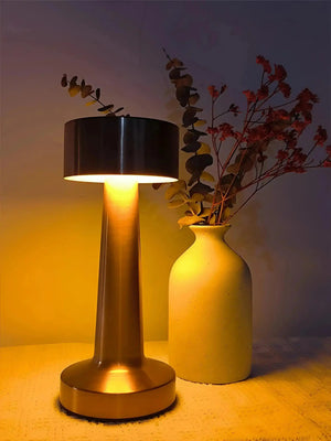 Table lamp with a black cylindrical shade and metallic base emitting warm light.